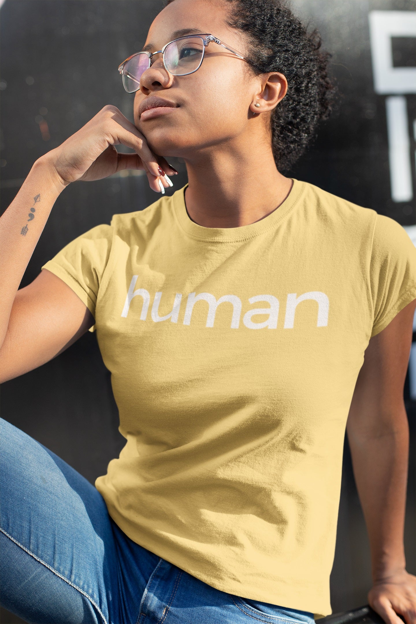 attractive woman sitting with her hand on her chin wearing yellow "human" t-shirt and denim pants