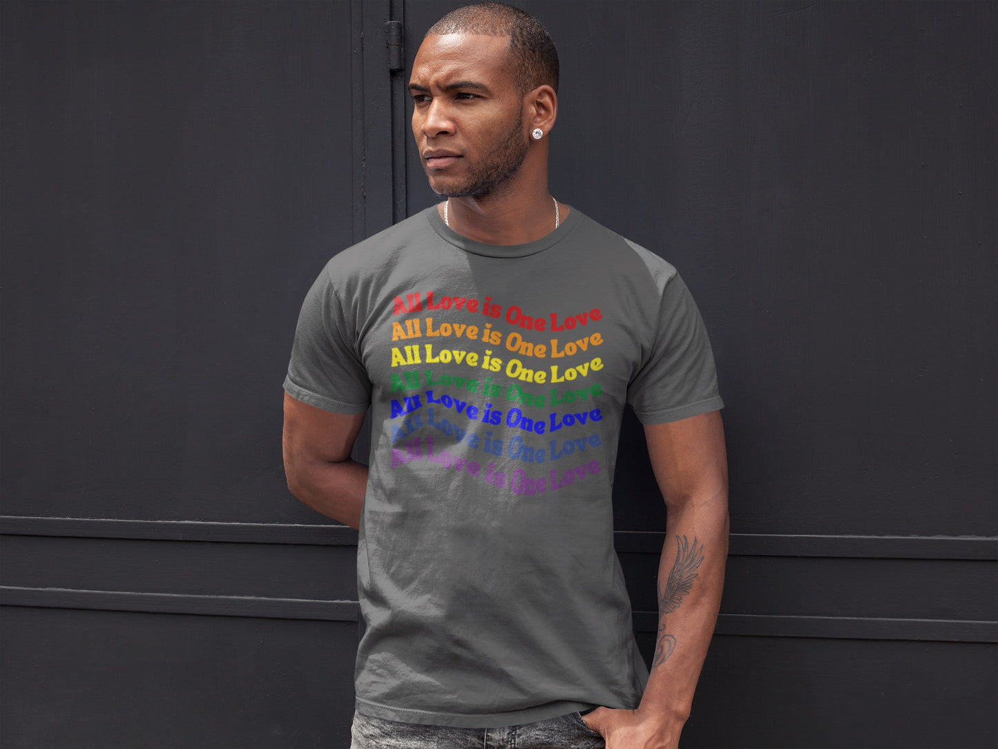 black man standing against a dark gray wall wearing an asphalt "all love is one love" graphic (repeated in rainbow colors) t-shirt