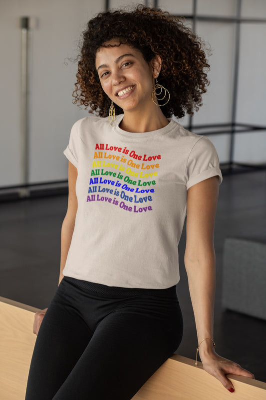 smiling curly haired attractive woman wearing a cream colored "all love is one love" graphic (repeated in rainbow colors) t-shirt and tight black pants sitting on a wooden partition wall