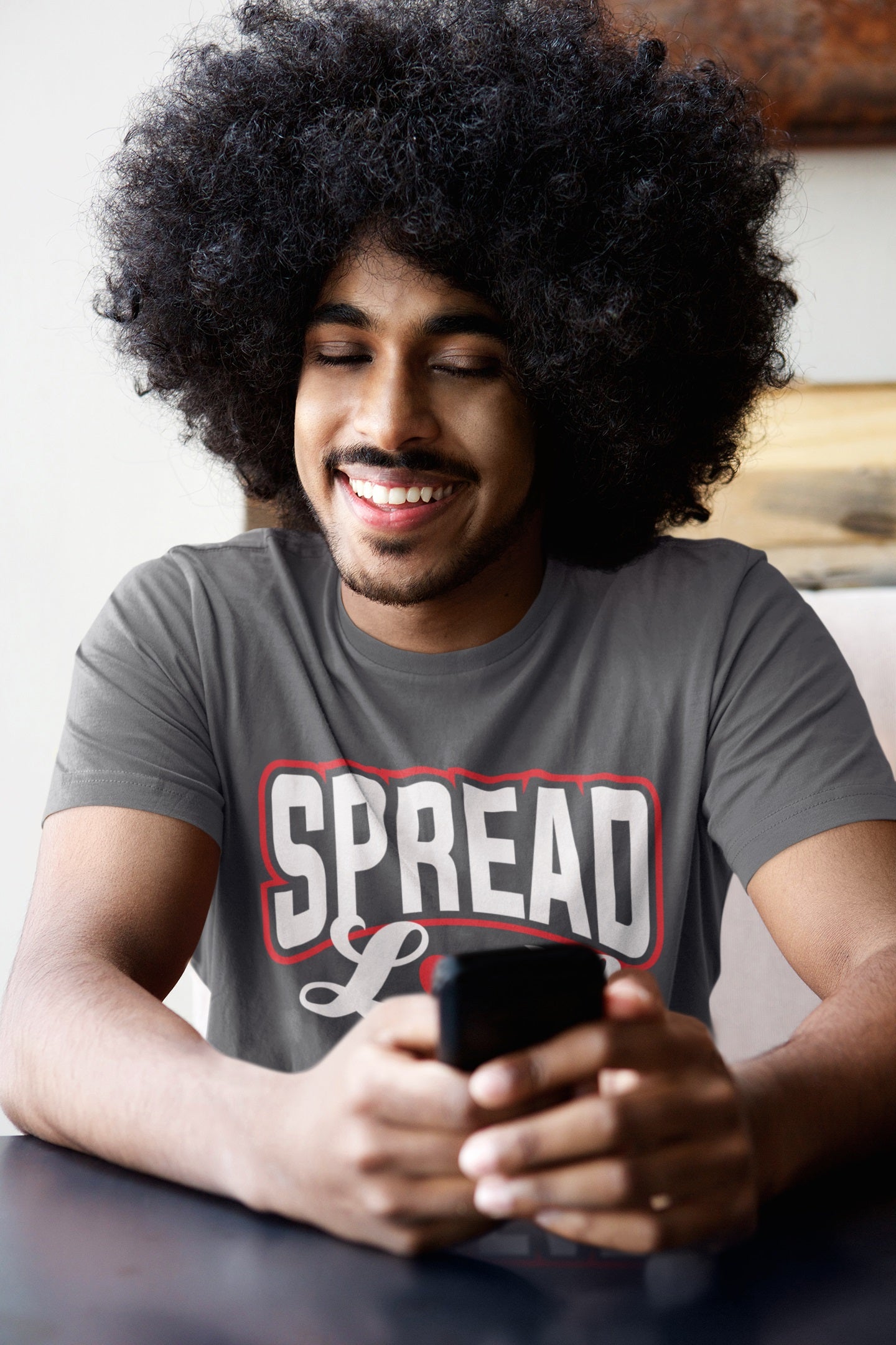 man with big curly afro wearing gray t-shirt with "spread love" graphic with red heart replacing the "o" sitting and looking at his cell phone