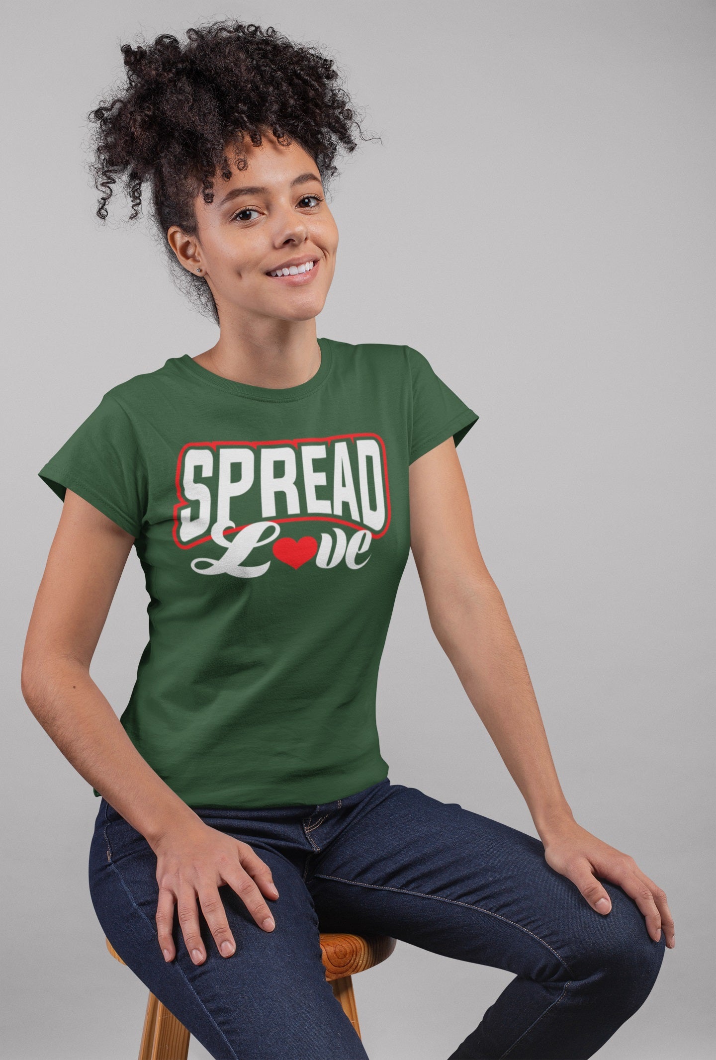 happy curly haired woman wearing forest green t-shirt with "spread love" graphic with red heart replacing the "o" while sitting on stool 