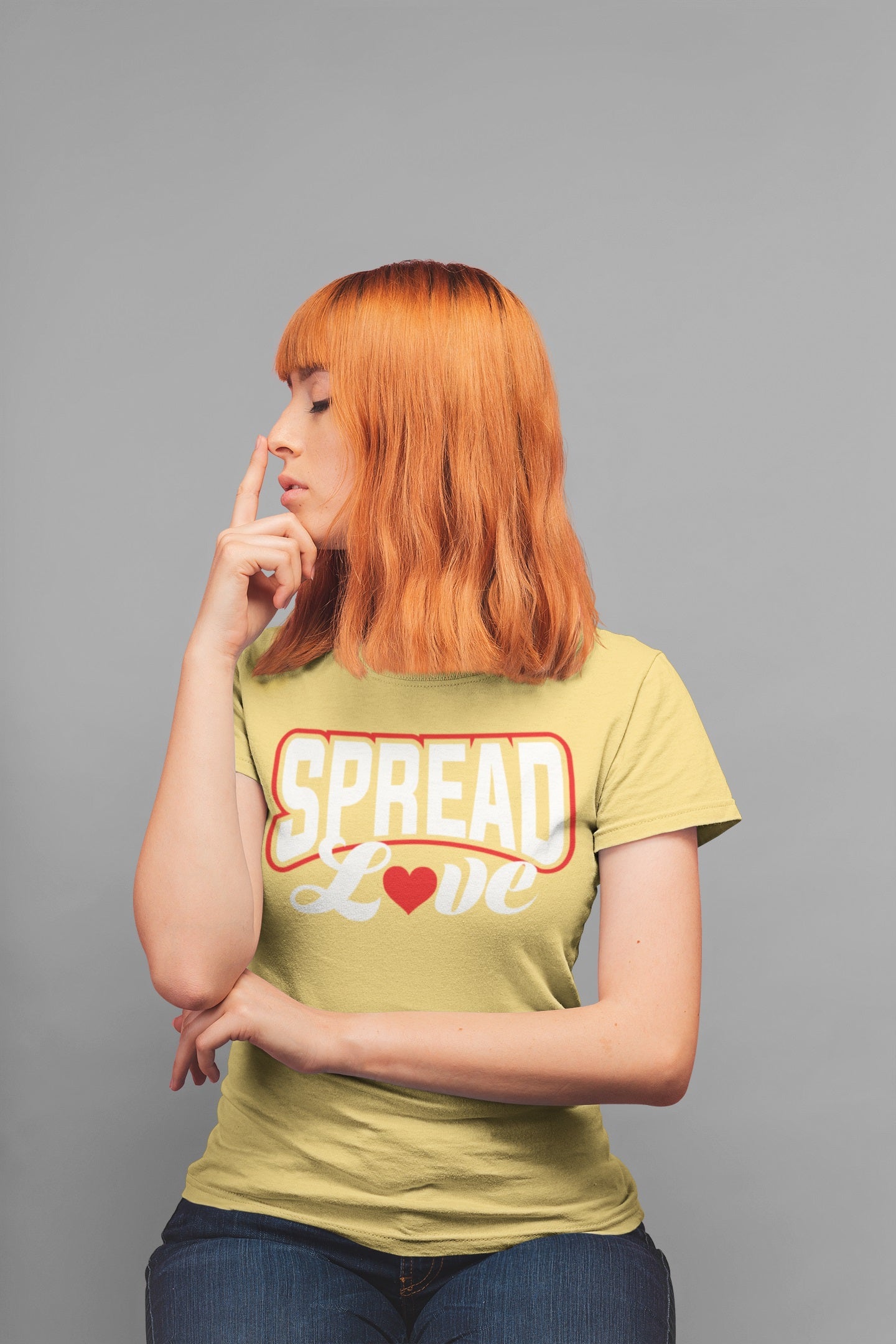 redhead woman sitting on a stool touching her nose with her index finger wearing a yellow t-shirt with "spread love" graphic with red heart replacing the "o" 