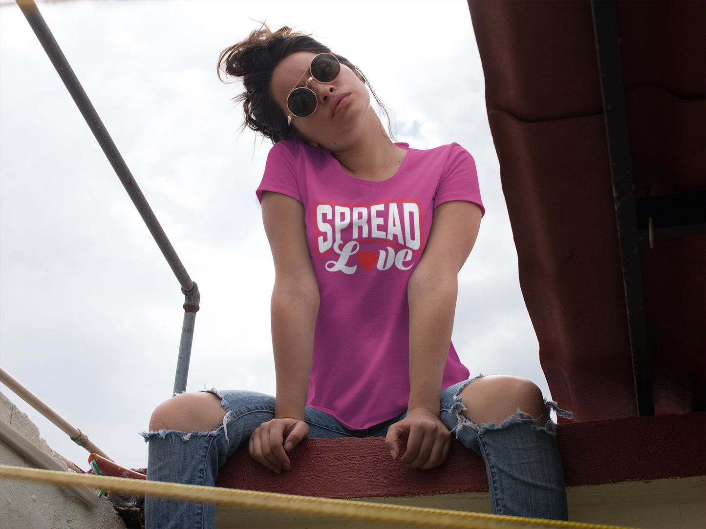 cute woman sitting on a wall wearing torn jeans and a berry t-shirt with "spread love" graphic with red heart replacing the "o" 