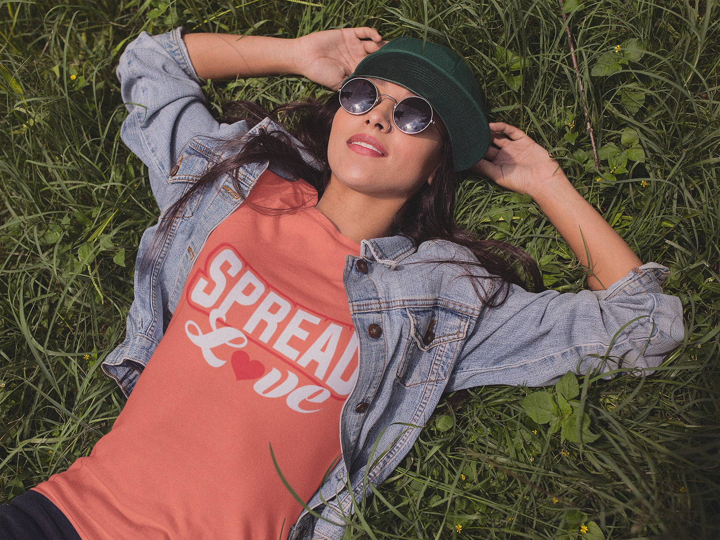 woman lying in the grass wearing a heather orange t-shirt with "spread love" graphic with red heart replacing the "o" under a denim jacket and also wearing sunglasses and a green cap