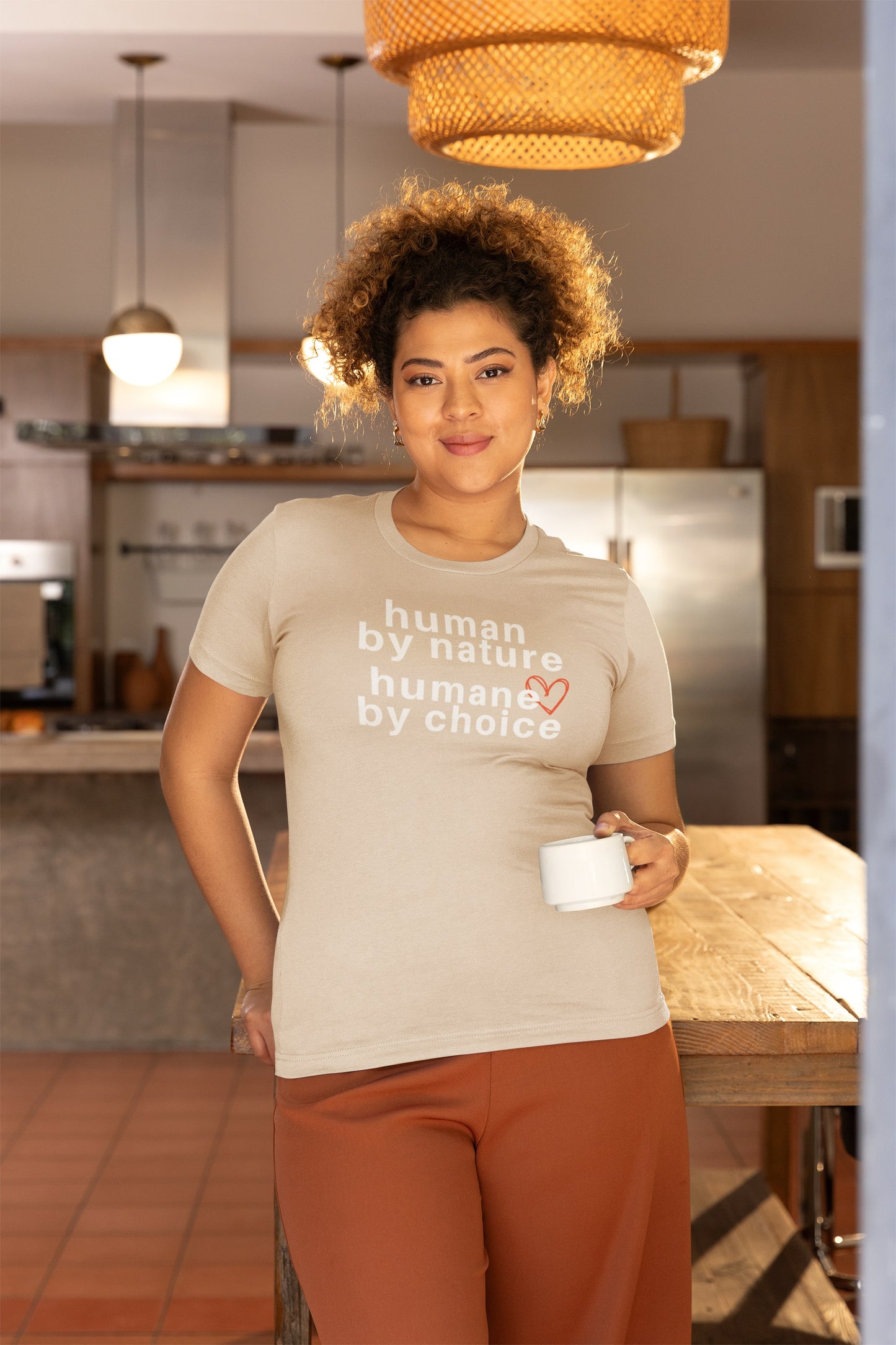 woman leaning against a wooden table wearing a cream t-shirt with " human by nature, humane by choice" graphic and holding a cup of coffee 