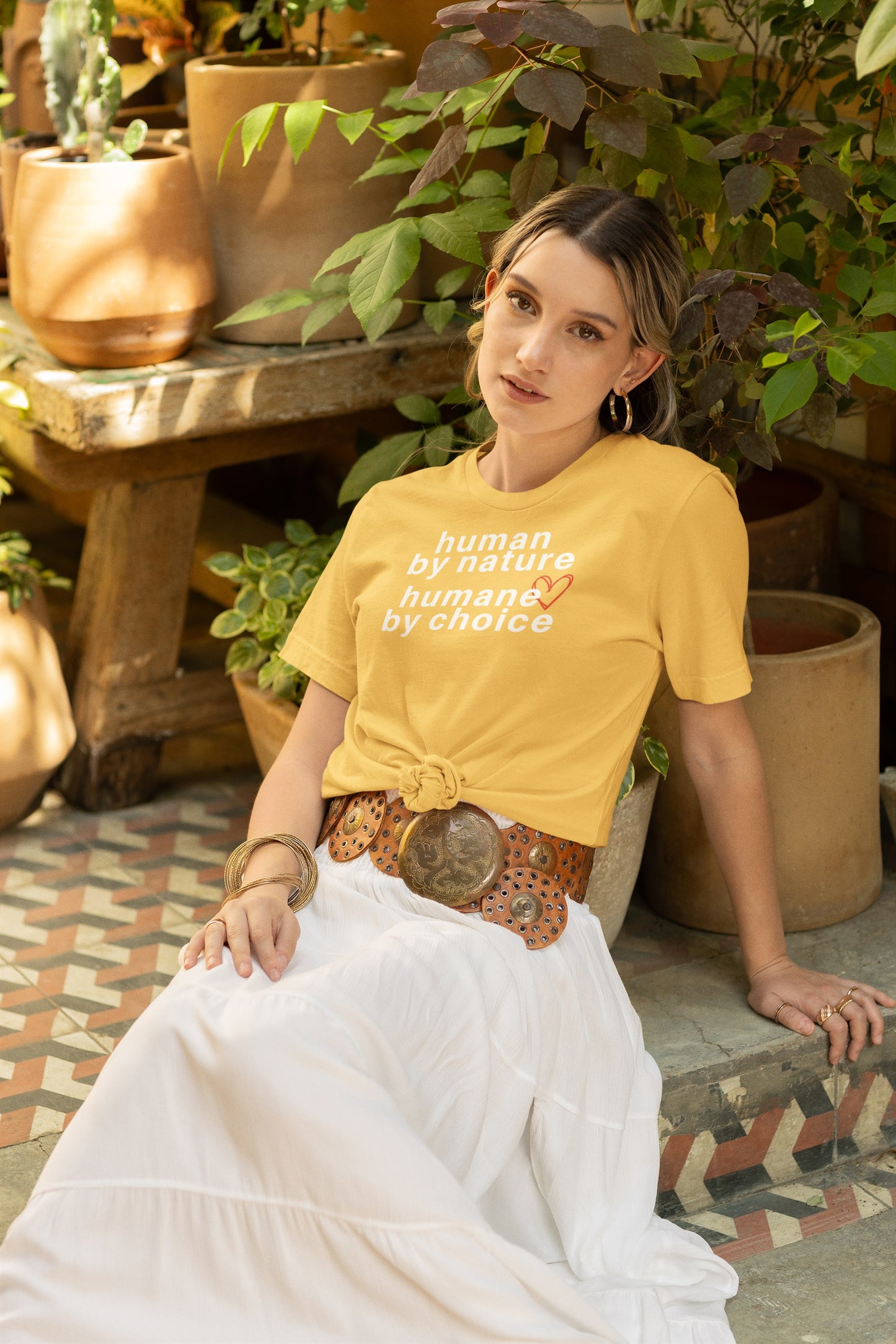 a woman posing by plant pots in a boho style outfit wearing yellow t-shirt with "human by nature, humane by choice" graphic.