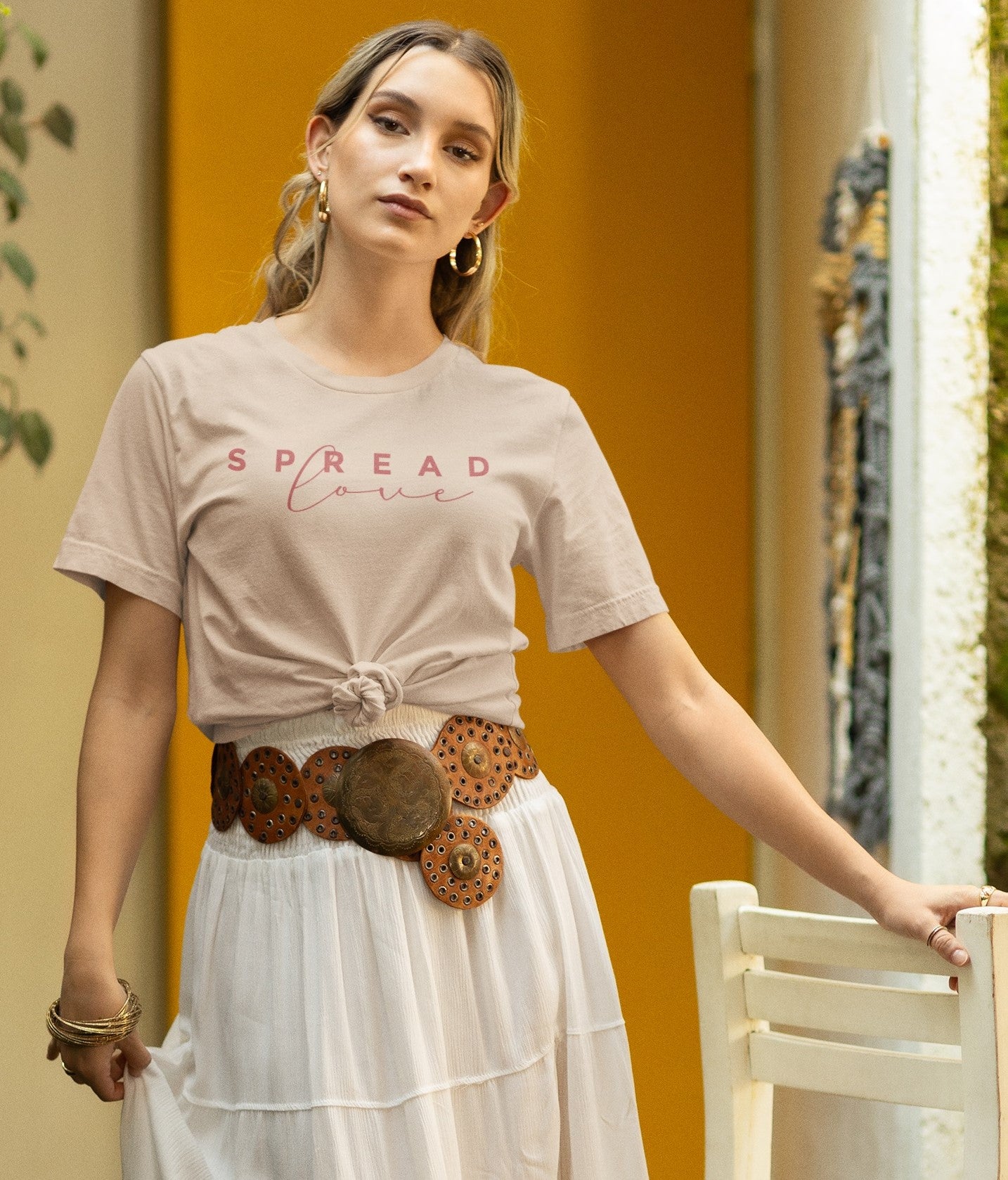 woman wearing a creak spread love t-shirt with her boho style outfit holding onto a white chair standing in a door way 